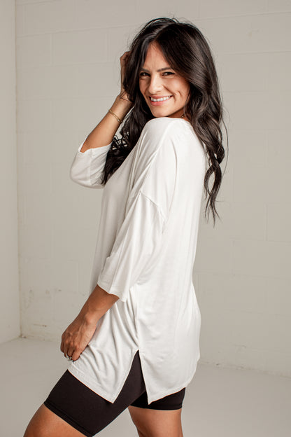 A woman modeling a white, oversized three-quarter sleeve tunic top with a high-low hem over black athletic shorts, standing against a light gray brick wall.
