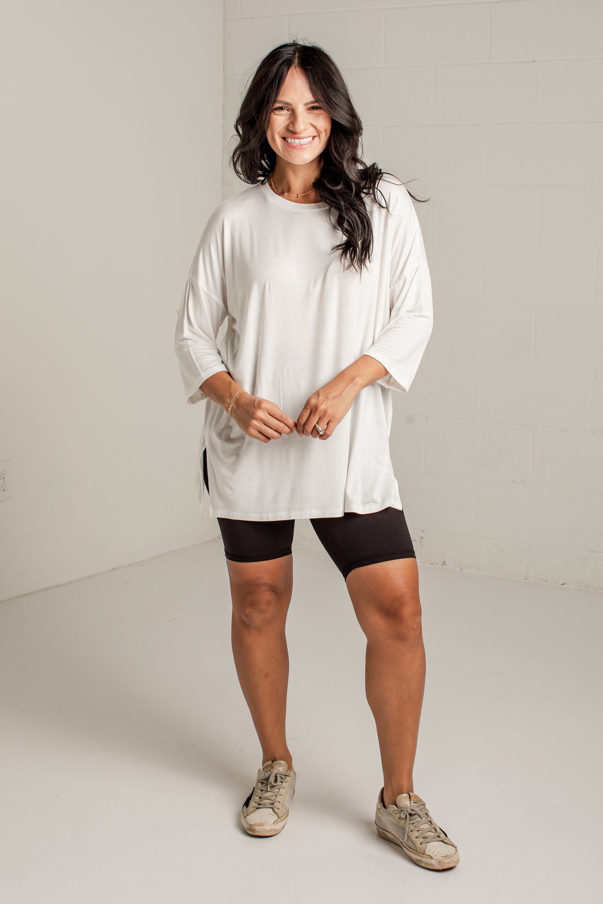 A woman modeling a loose-fitting off-white three-quarter sleeve tunic top, paired with black bike shorts and beige sneakers, standing in a bright, neutral studio setting.
