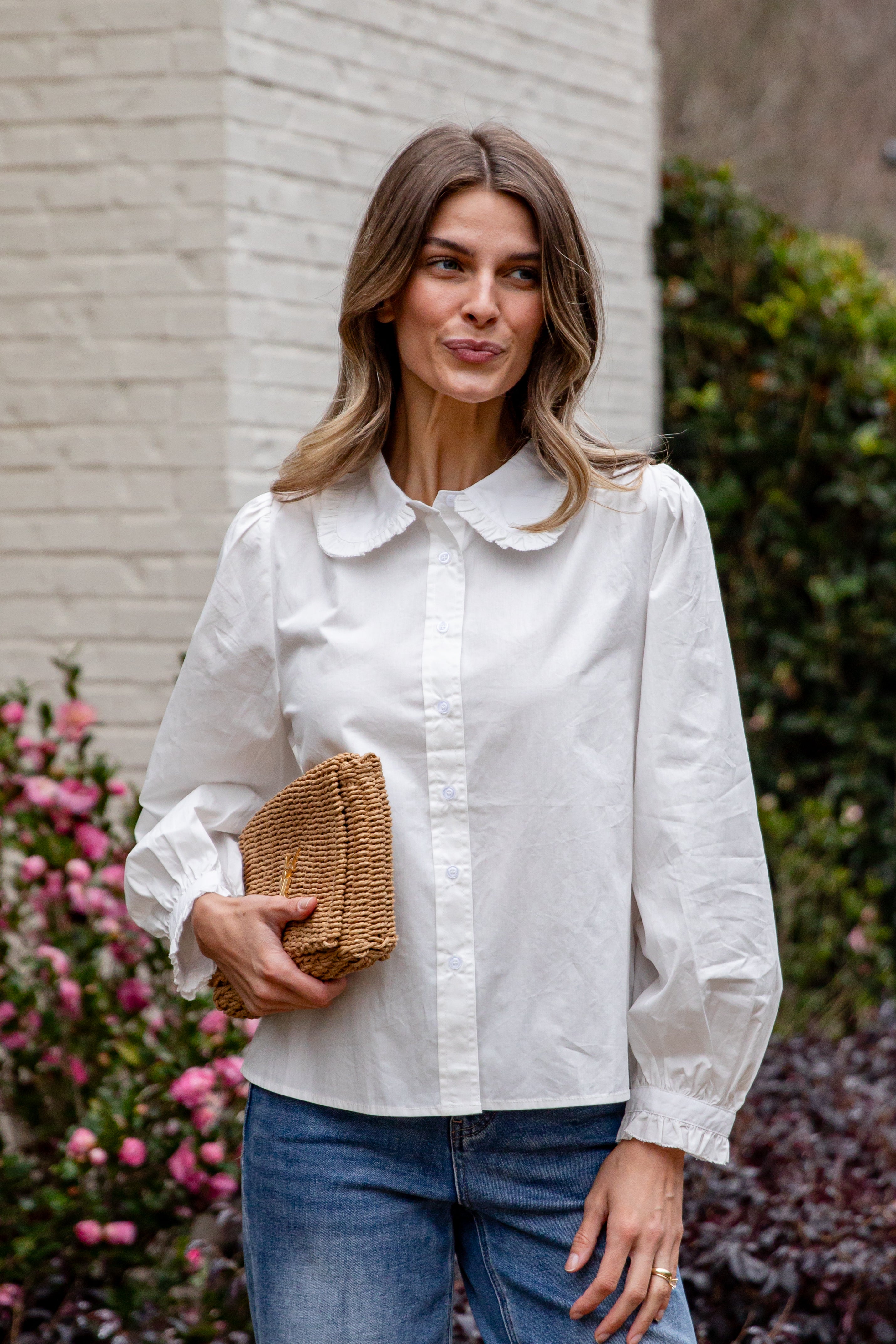 Woman wearing a white blouse and blue jeans, holding a woven clutch against a brick wall and floral background.