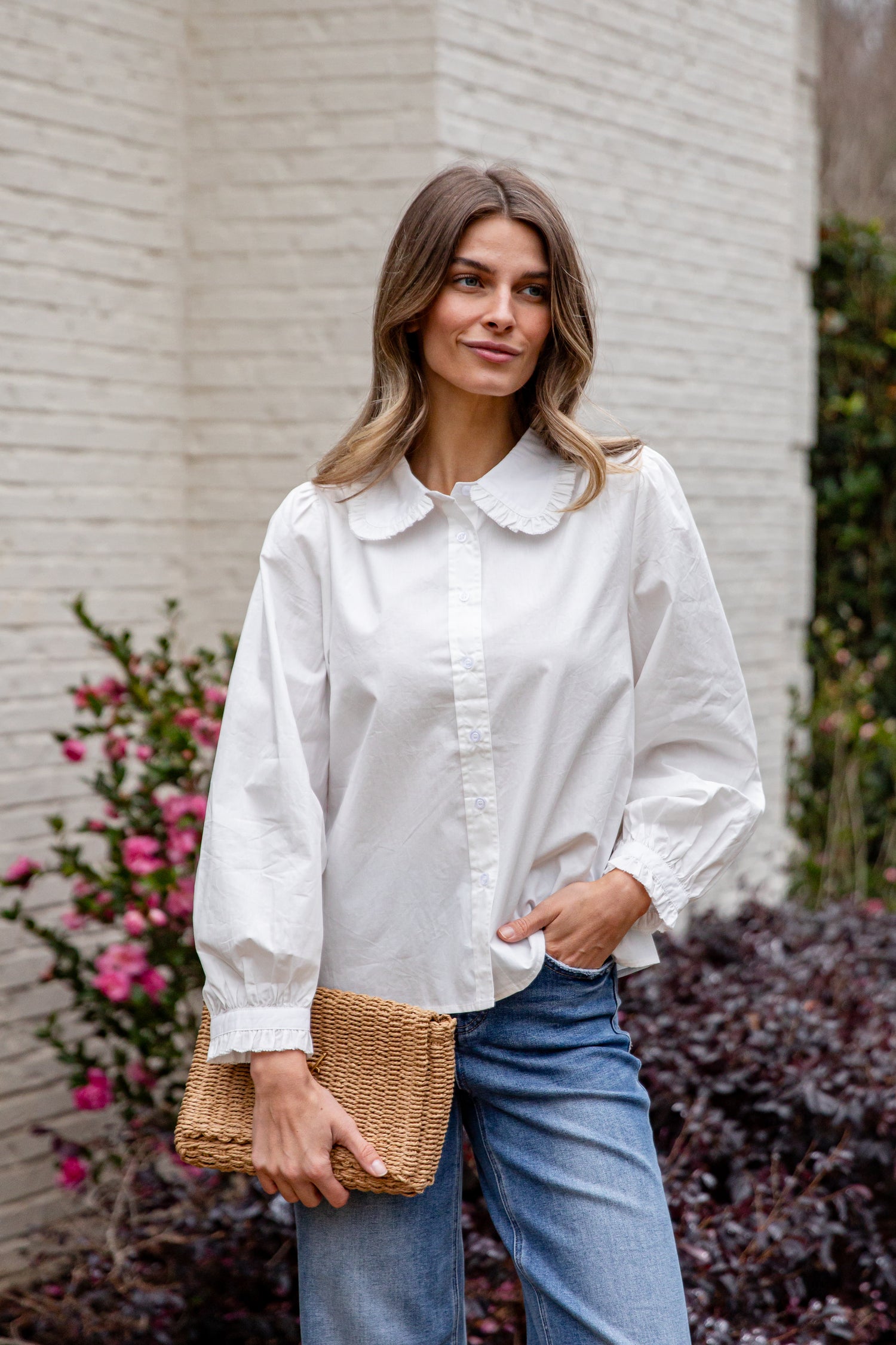 Woman wearing a white blouse and blue jeans, holding a woven clutch, standing against a light-colored wall with plants.