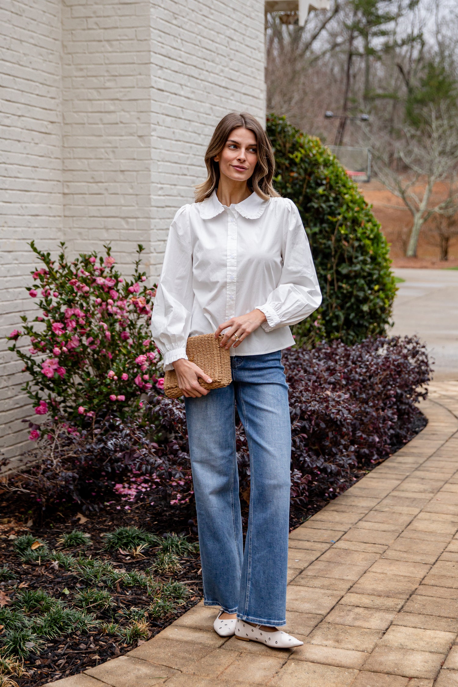 Woman in a white blouse and blue jeans standing outdoors near a building with plants.