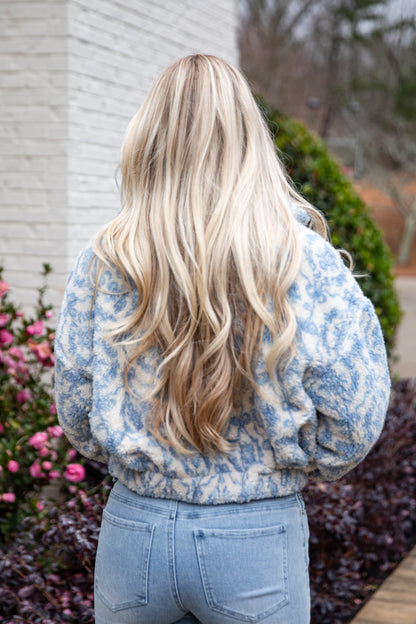 Person with long blonde hair wearing a blue floral sweater and jeans, standing outdoors.