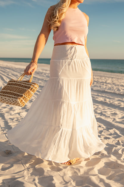 Woman in a pink top and white skirt holding a straw bag on a sandy beach.