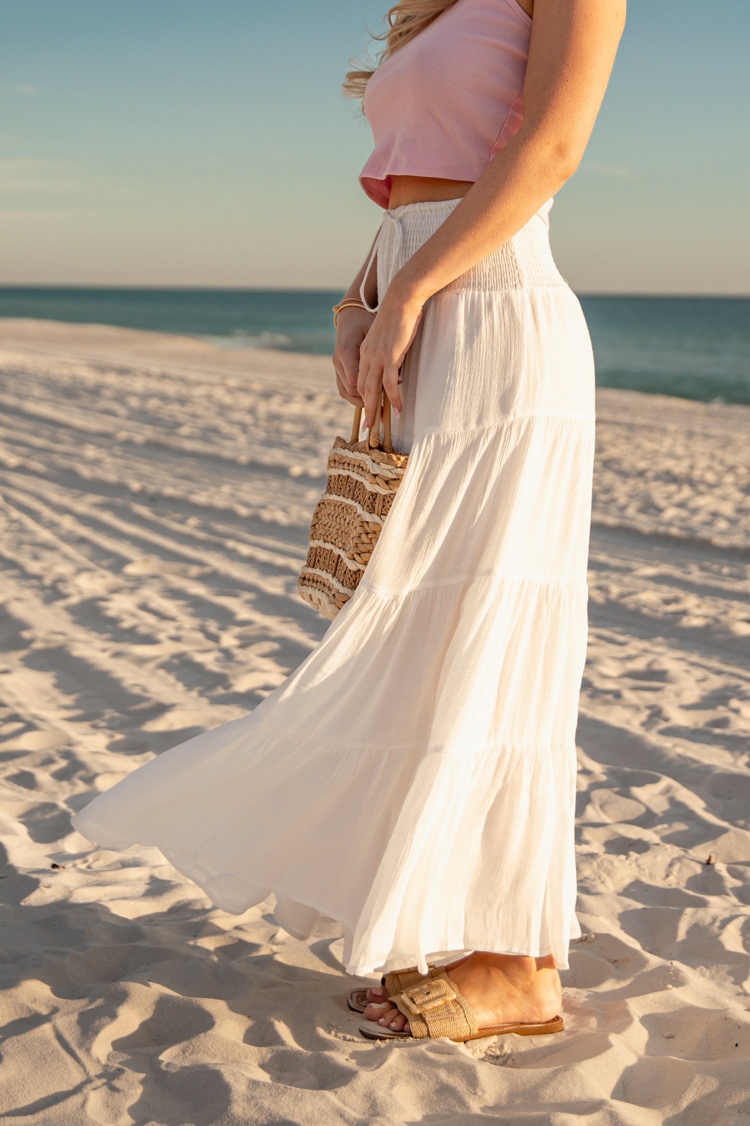 Woman in a white dress and pink top standing on a sandy beach with ocean view.