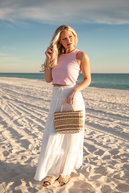 Woman on a beach holding a woven bag, wearing a pink top and white skirt.