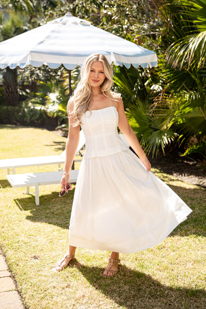 Woman in a white dress standing outdoors under a white umbrella with greenery in the background