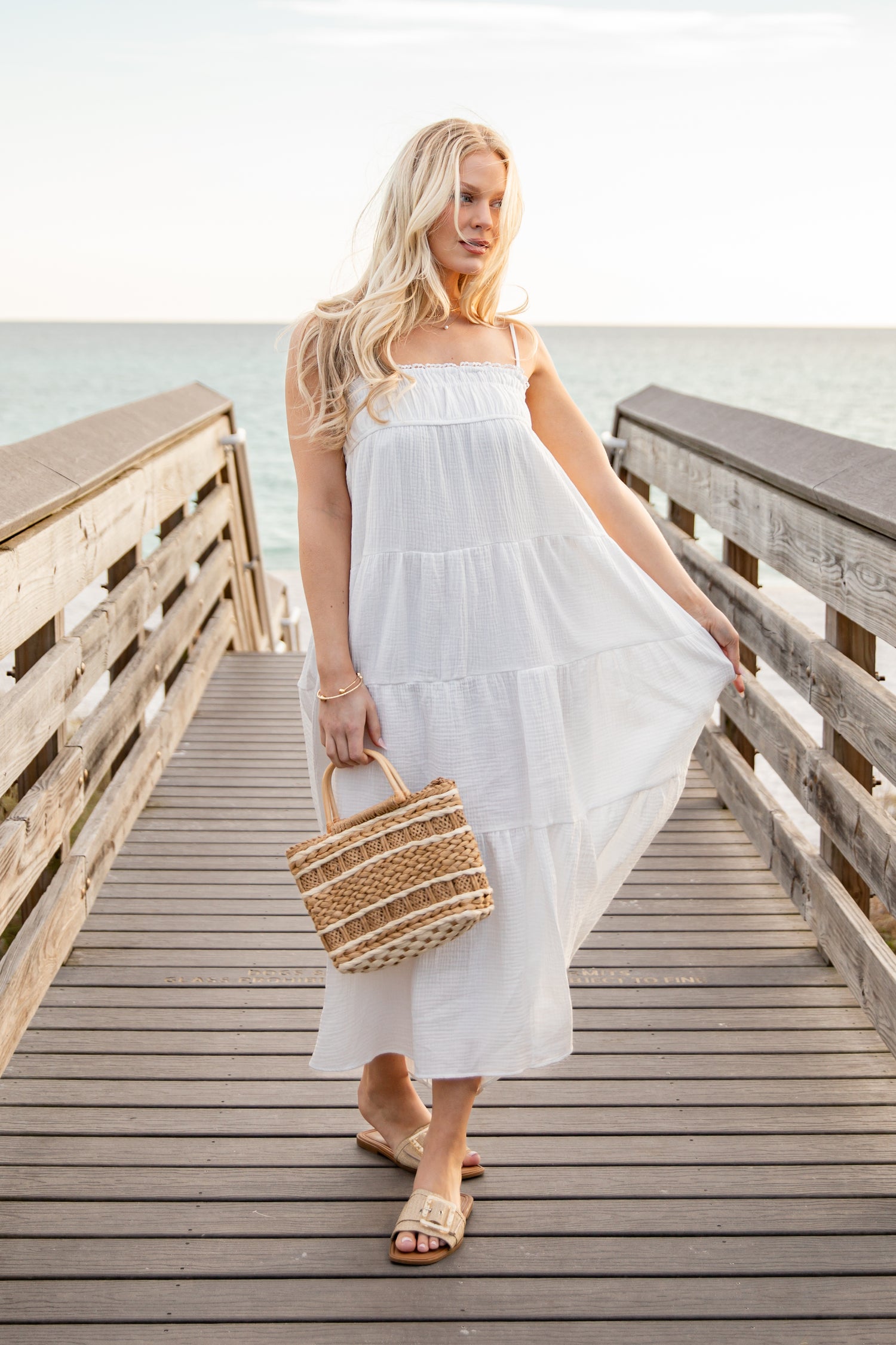 Woman in a white dress holding a straw bag on a wooden dock by the water.