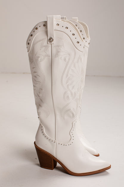 A single white faux-leather cowboy boot with embossed floral detailing, silver studs, and a brown stacked block heel, photographed on a light gray studio background.