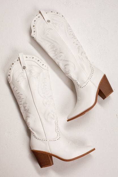 A pair of white western cowboy boots with brown stacked block heels, shown from a top-down angle on a light off-white background.