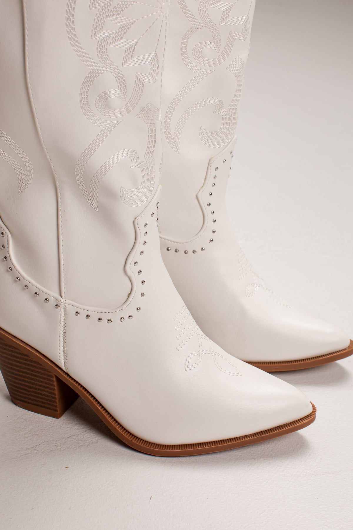 A pair of white faux leather cowboy boots with decorative western embroidery, studded trim, and brown stacked block heels, shown against a light gray studio background.