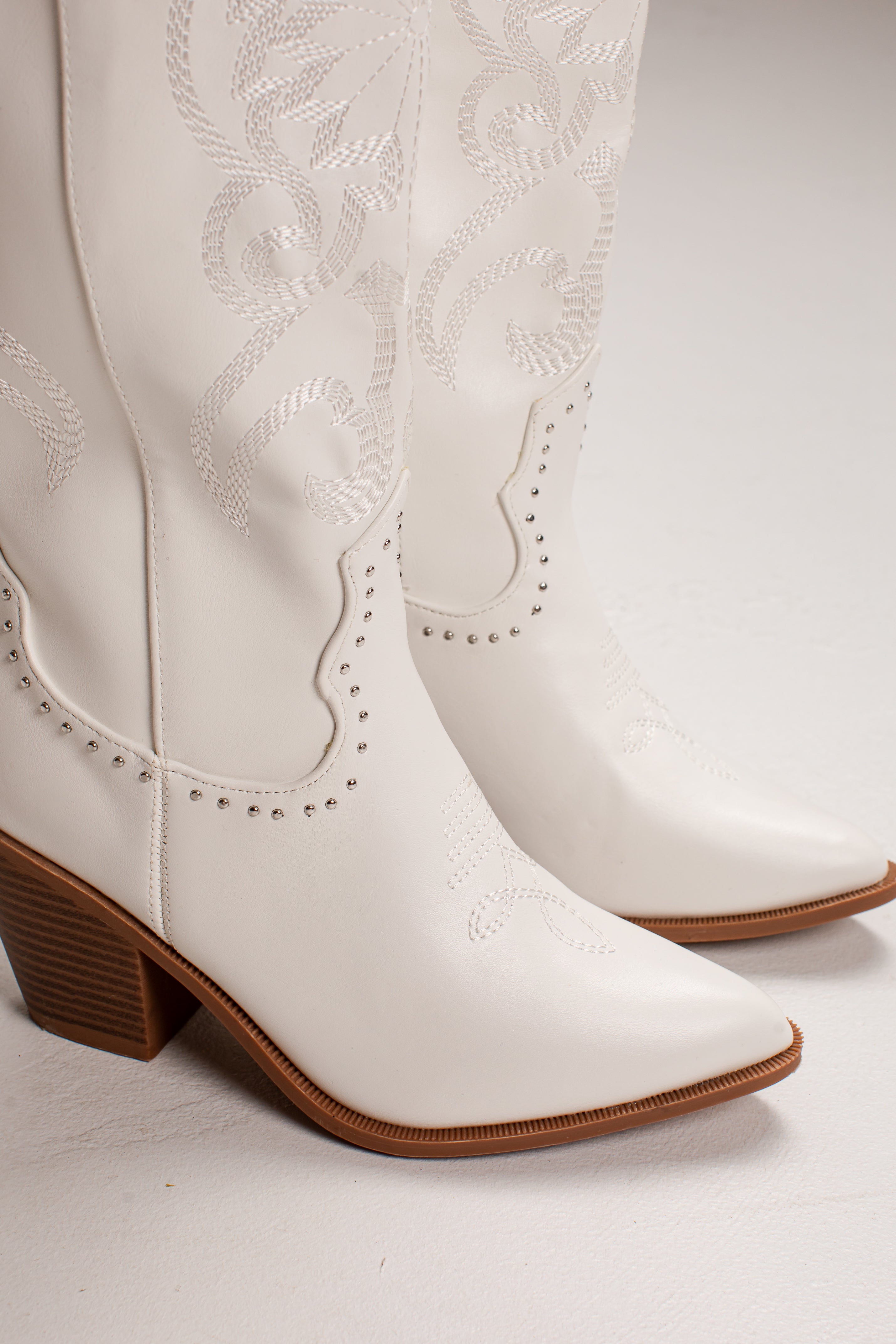 A pair of white faux leather cowboy boots with decorative western embroidery, studded trim, and brown stacked block heels, shown against a light gray studio background.
