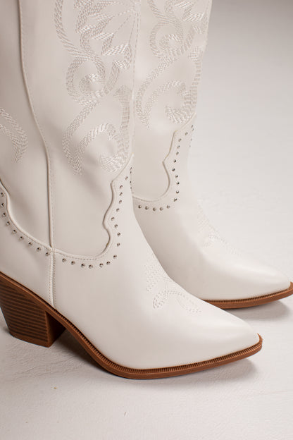 A pair of white faux leather cowboy boots with decorative western embroidery, studded trim, and brown stacked block heels, shown against a light gray studio background.