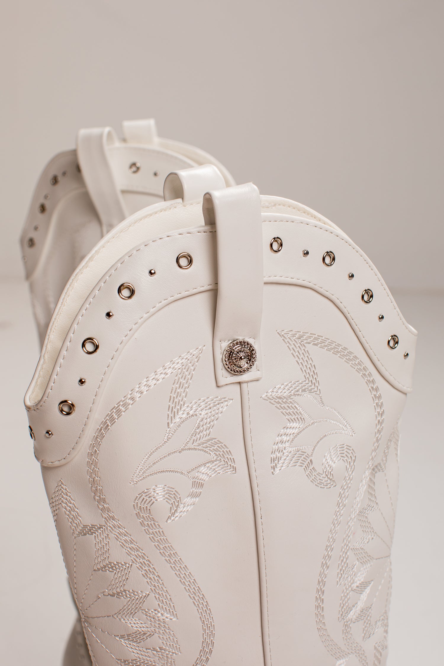 Close-up of a white western boot with silver-toned studs, decorative stitching, and a central concho button, shown against a neutral light gray background.