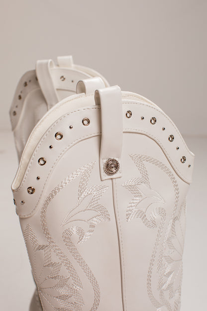 Close-up of a white western boot with silver-toned studs, decorative stitching, and a central concho button, shown against a neutral light gray background.