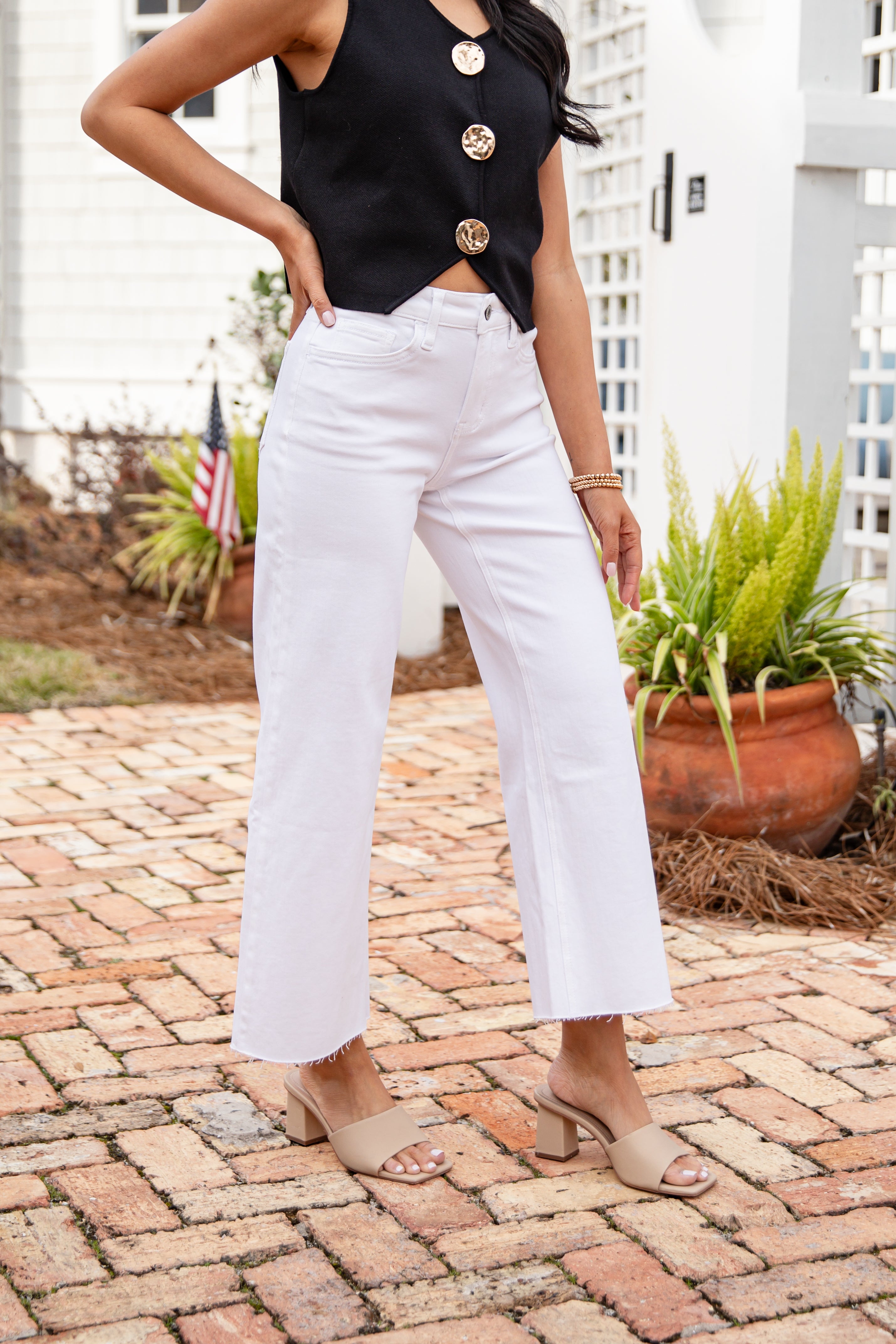 Person wearing a black top with gold buttons and white pants standing on a brick patio.