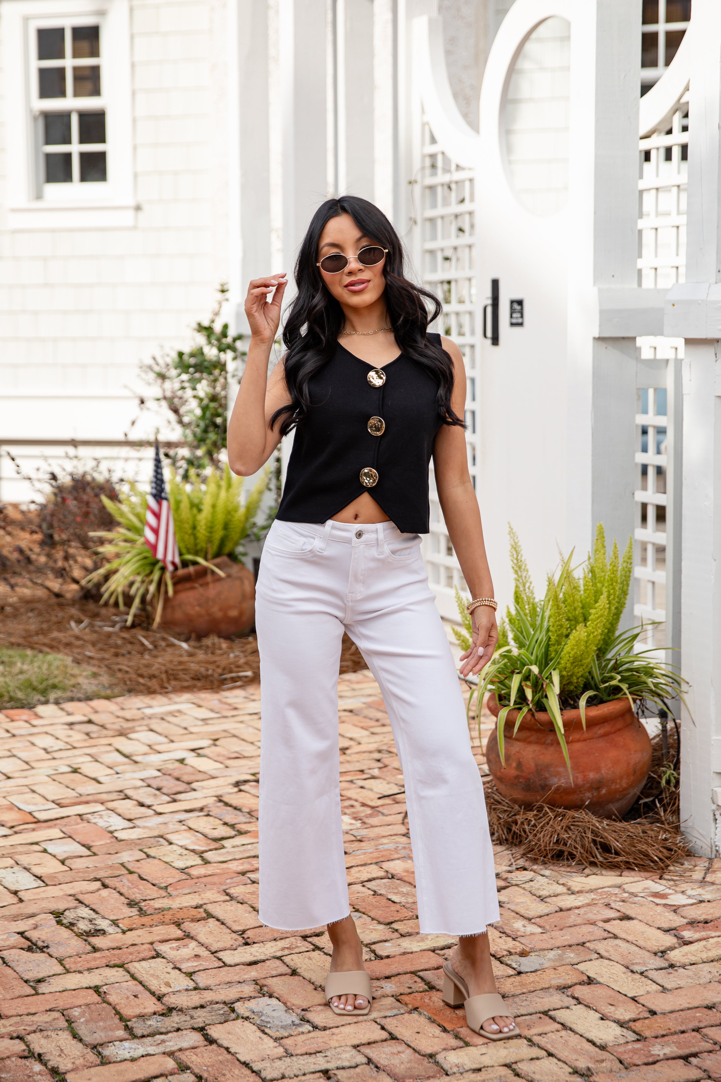 Woman in black top and white pants standing on a brick patio with plants and a white building in the background.