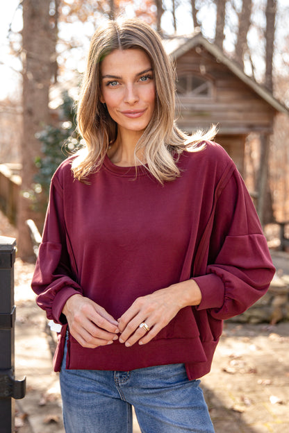 Woman wearing a burgundy sweater and blue jeans standing outdoors with trees in the background
