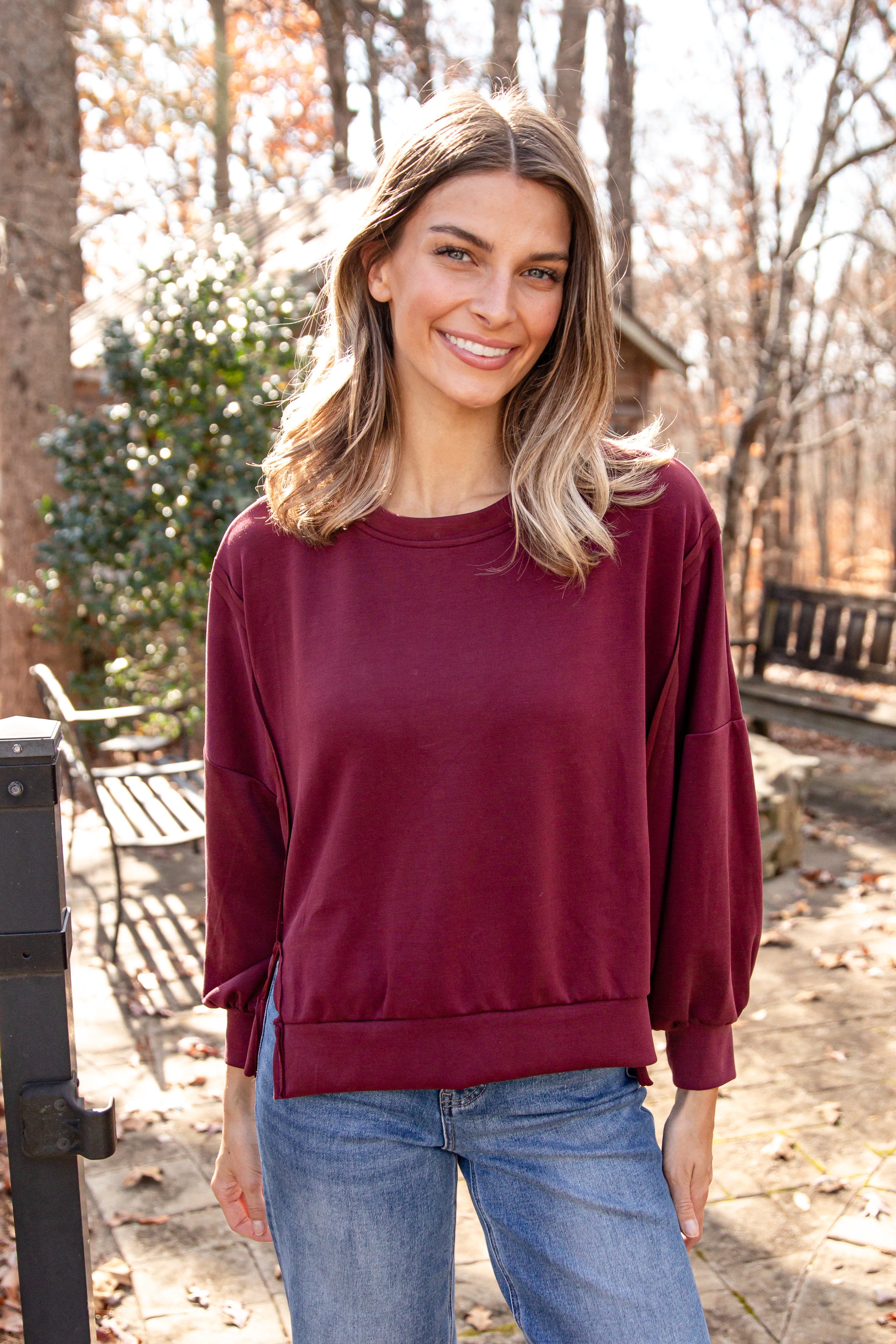 Woman wearing a maroon sweater and blue jeans standing outdoors with trees and a bench in the background.