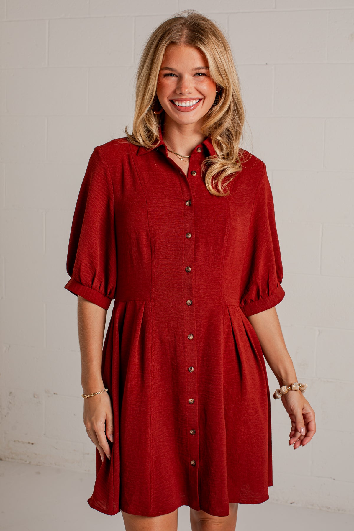 A smiling blonde woman wearing a textured red button-down shirt dress with puff sleeves, standing against a white painted brick wall.