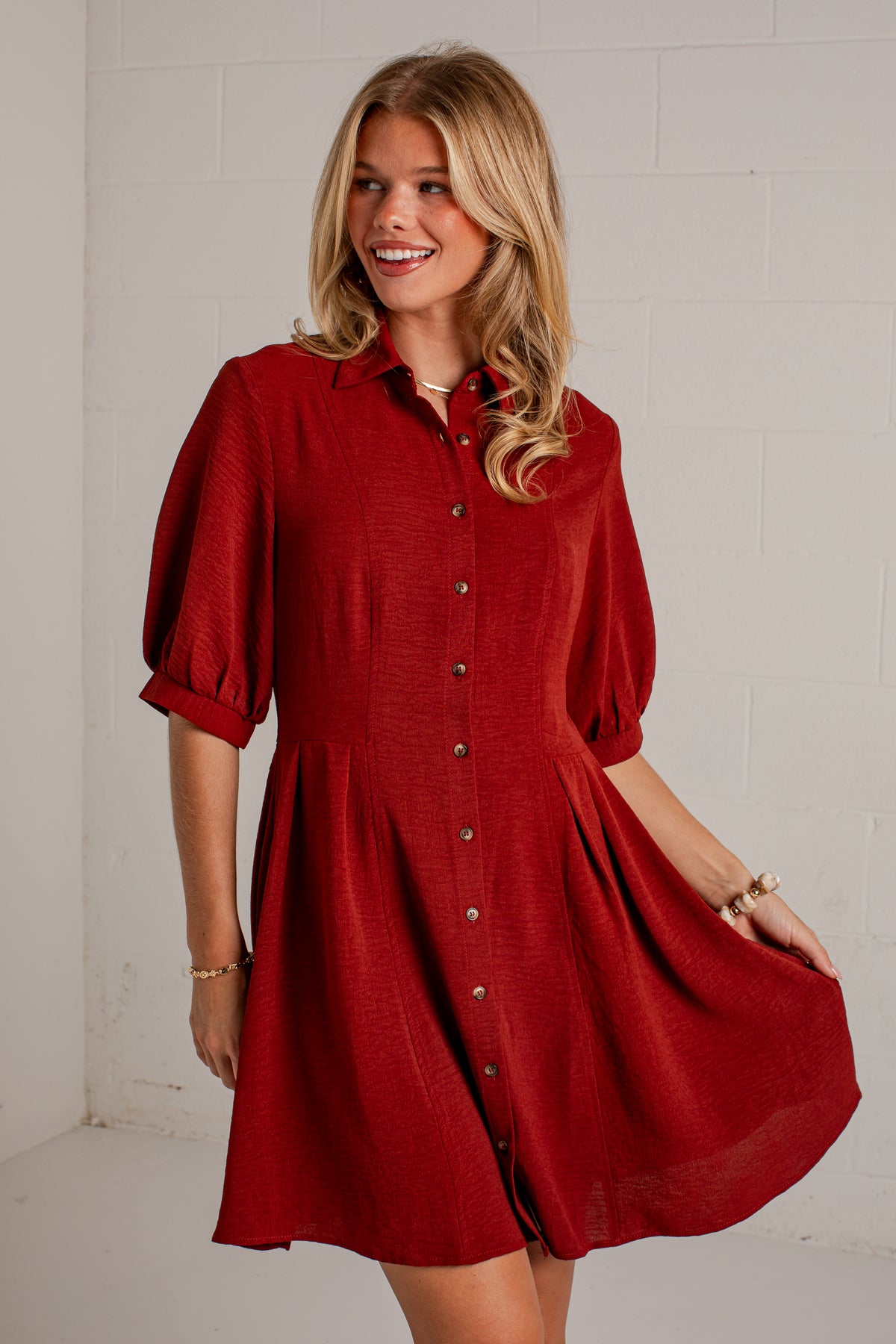 A woman wearing a red button-front shirt dress with puff sleeves and a flared skirt, standing against a white painted brick wall.