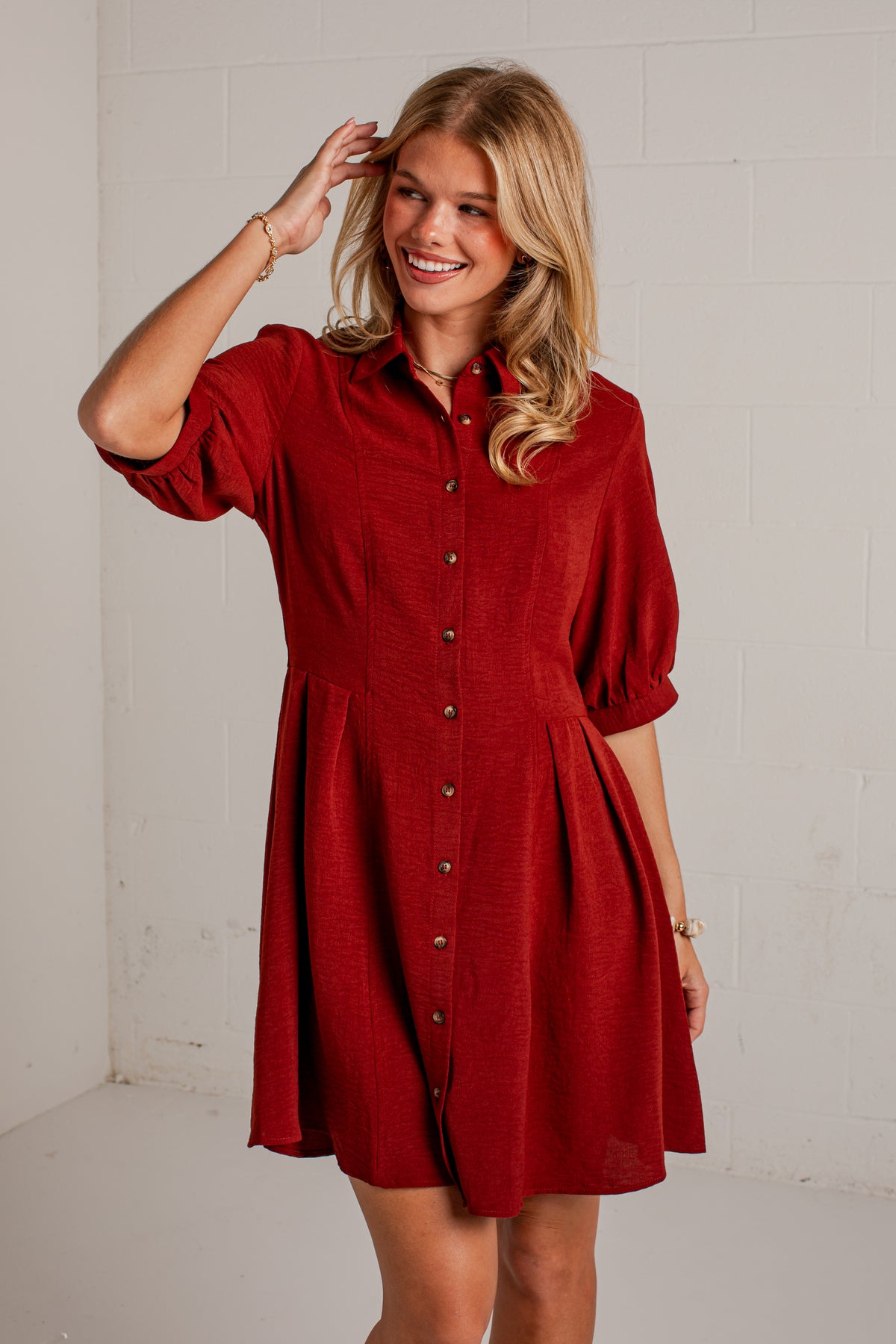 A woman modeling a red button-down shirt dress with puff sleeves and a flared skirt, standing against a white brick wall.