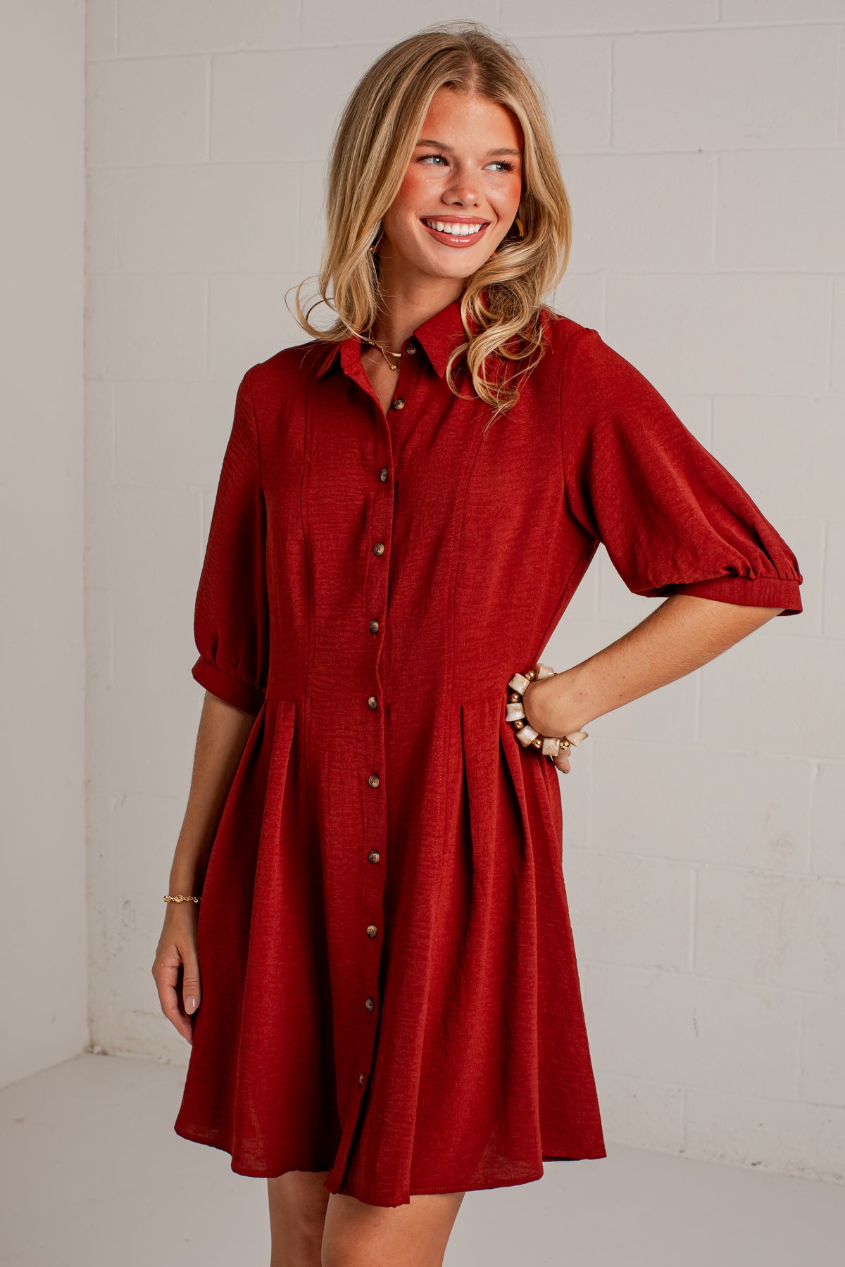 A woman modeling a red button-down shirt dress with puff sleeves and a flared skirt, standing against a white brick wall.