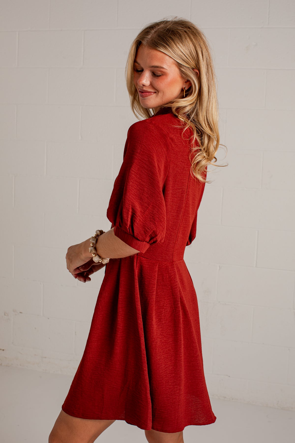 A woman wearing a red textured short puff-sleeve dress, photographed from a side-back angle against a white painted brick wall.