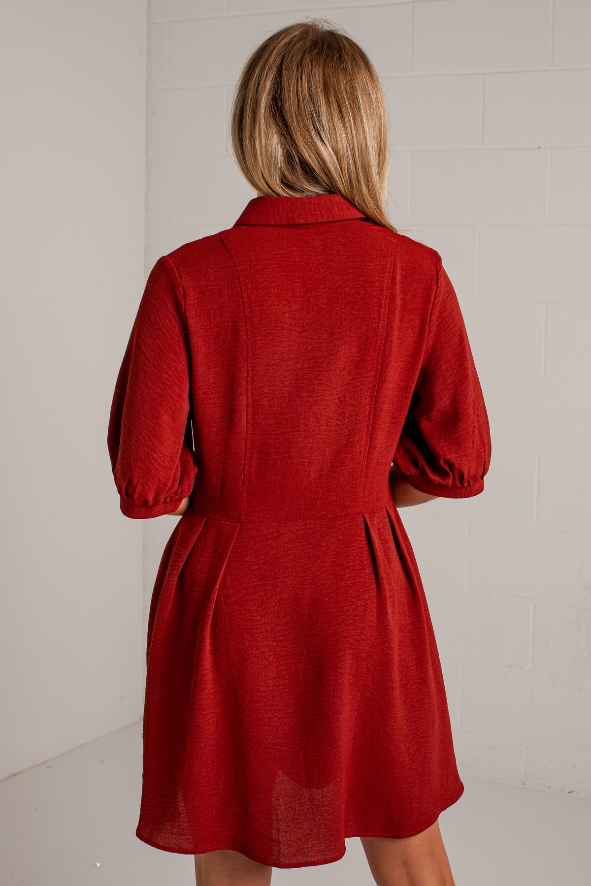A woman viewed from behind wearing a red short-sleeve collared dress with a gathered waist and flared skirt, standing against a white wall.