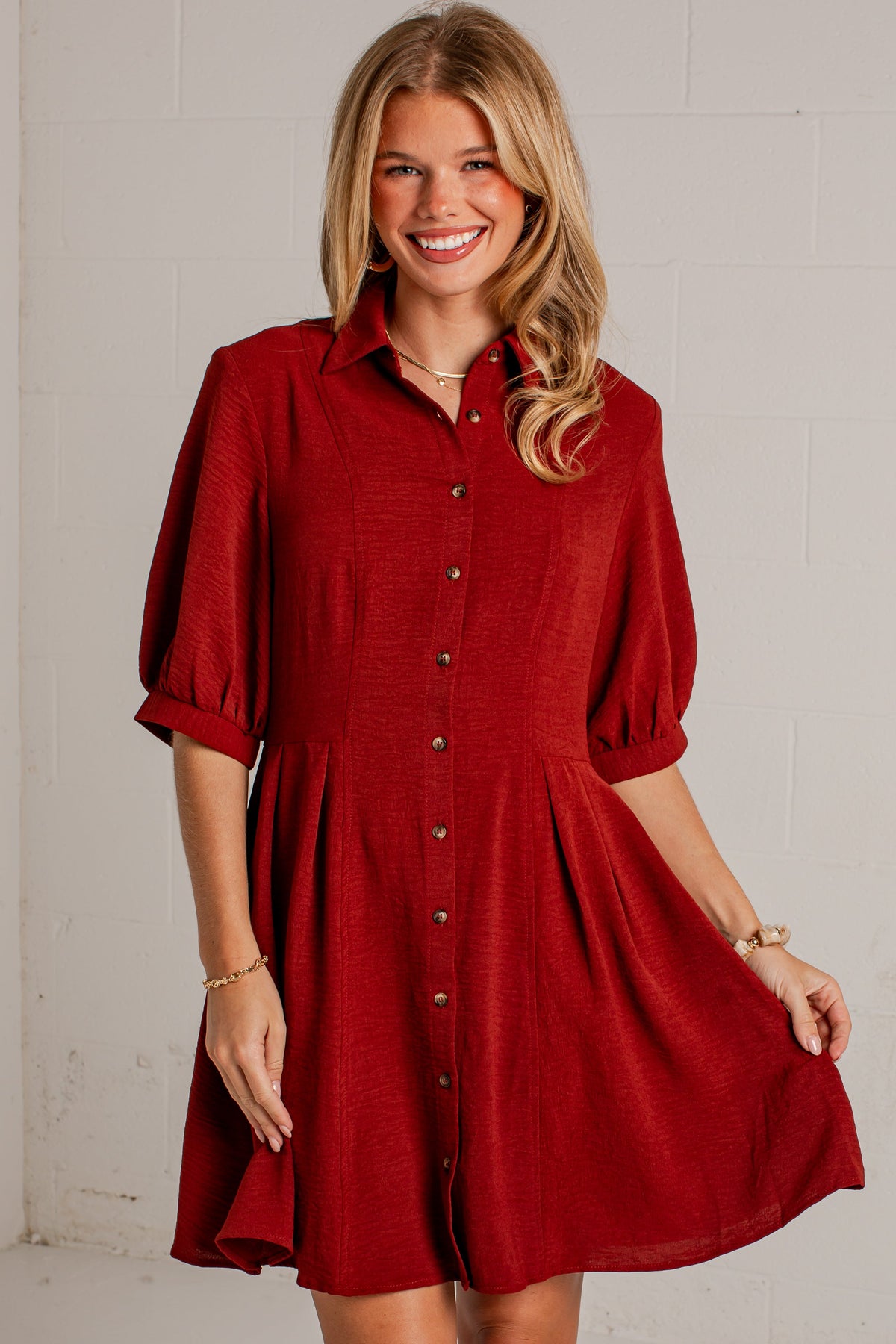 A smiling blonde woman models a short red button-up shirt dress with puff sleeves and a flared skirt, standing against a white painted brick wall.