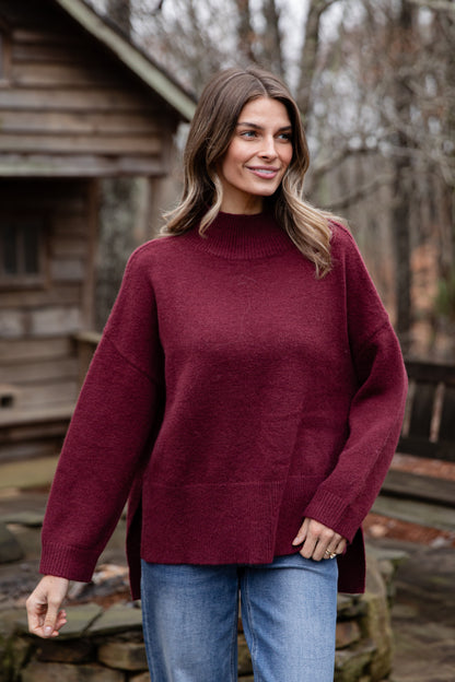 Woman wearing a maroon sweater and blue jeans standing outdoors with wooden structures and trees in the background.