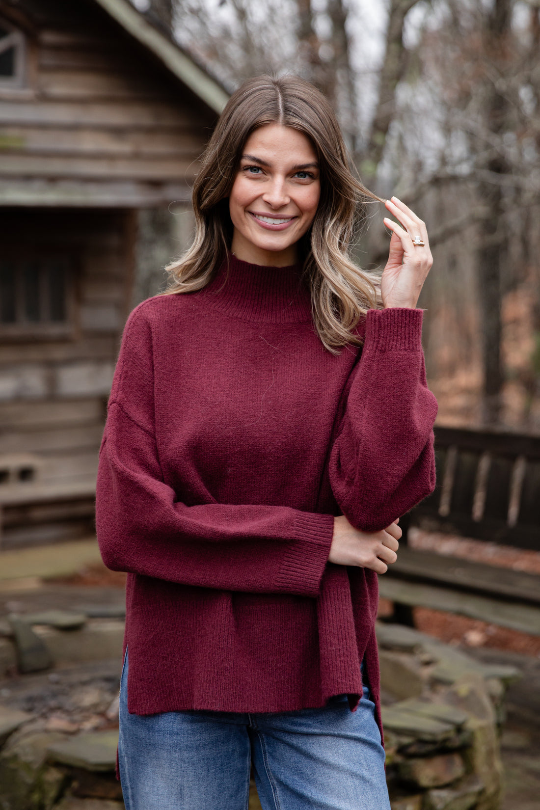 Woman wearing a maroon sweater and blue jeans standing outdoors near a wooden cabin.