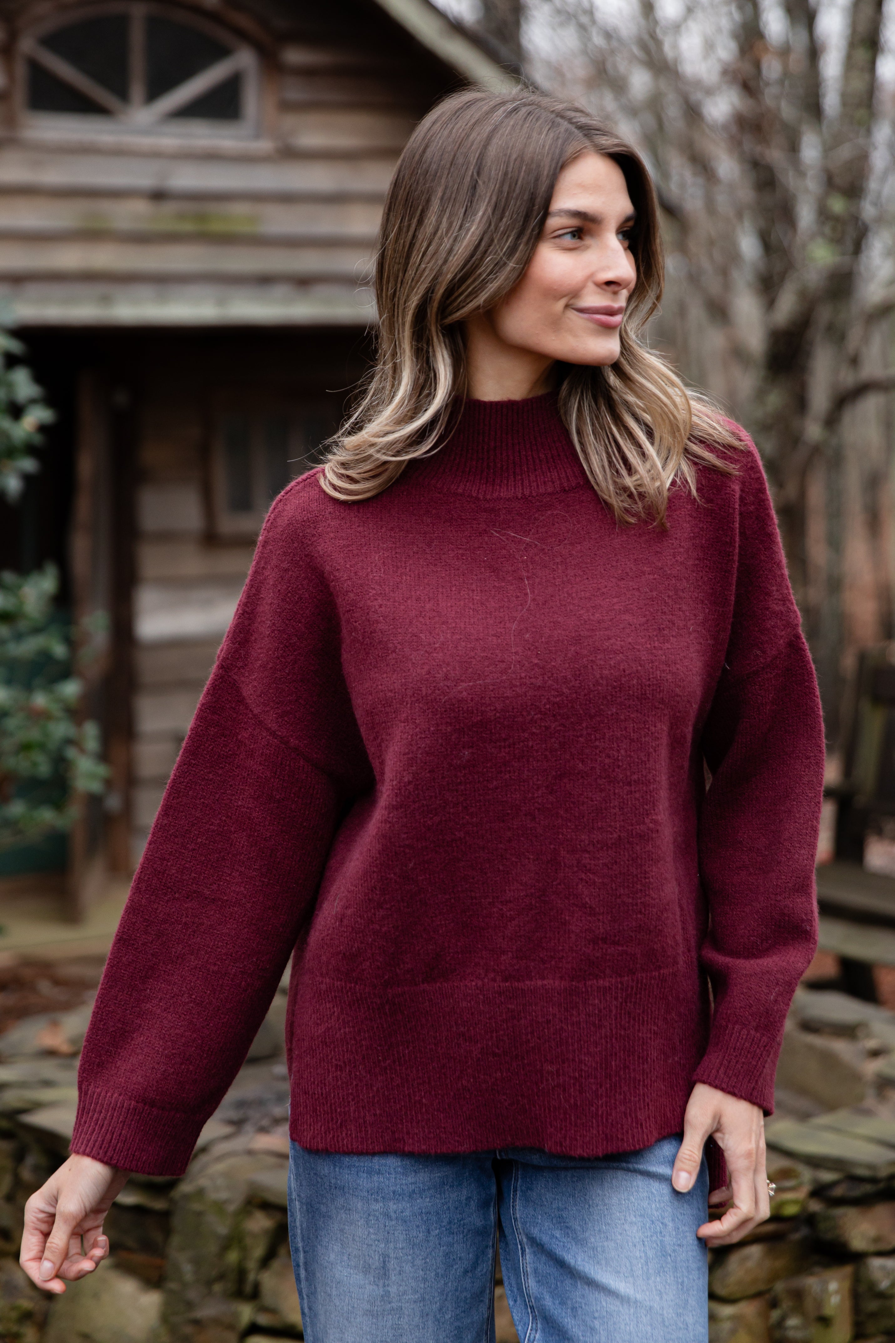 Woman wearing a maroon sweater and blue jeans standing outdoors.