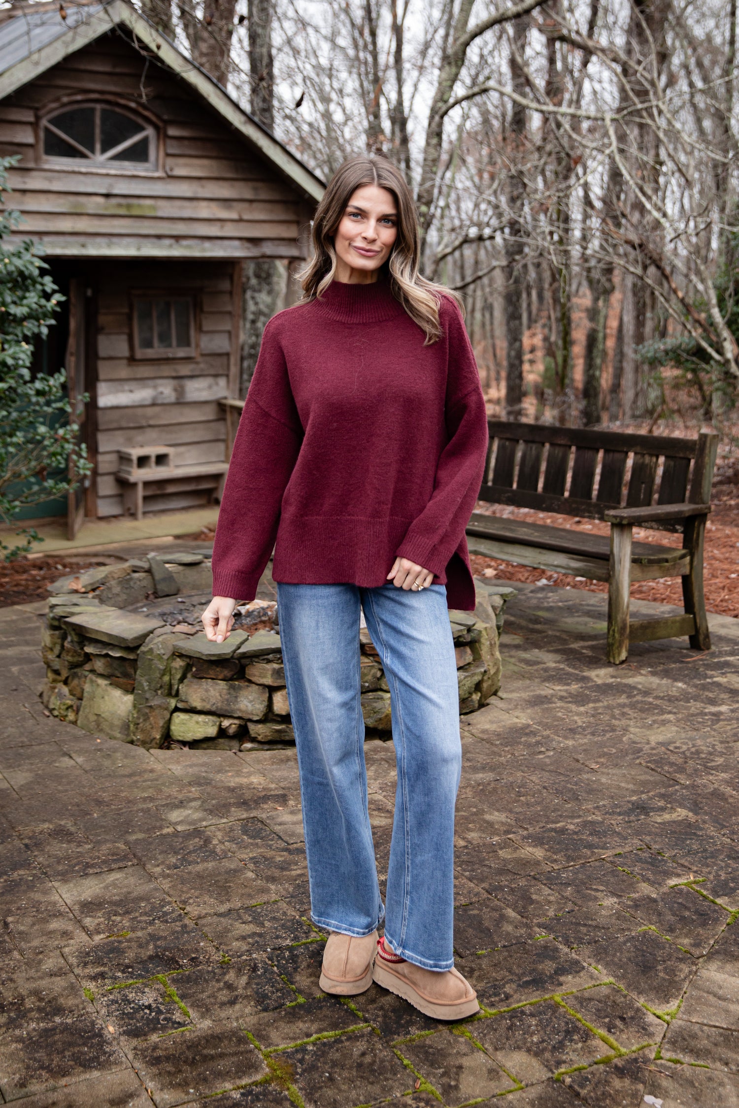 Woman wearing a burgundy sweater and blue jeans standing in front of a wooden cabin.