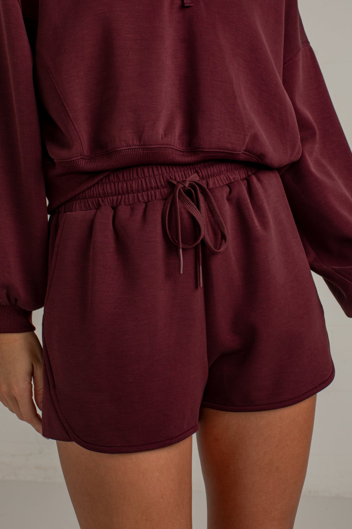 A close-up of a person wearing a matching burgundy lounge set, featuring a cropped sweatshirt and drawstring shorts, against a neutral light gray background.