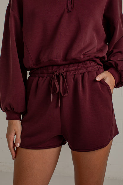 A person wearing a matching burgundy lounge set consisting of a cropped sweatshirt and drawstring shorts, shown from the chest down against a light neutral wall.