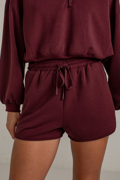 A close-up of a person wearing a matching burgundy lounge set, featuring a cropped long-sleeve top and high-waisted shorts with an elastic drawstring waist.