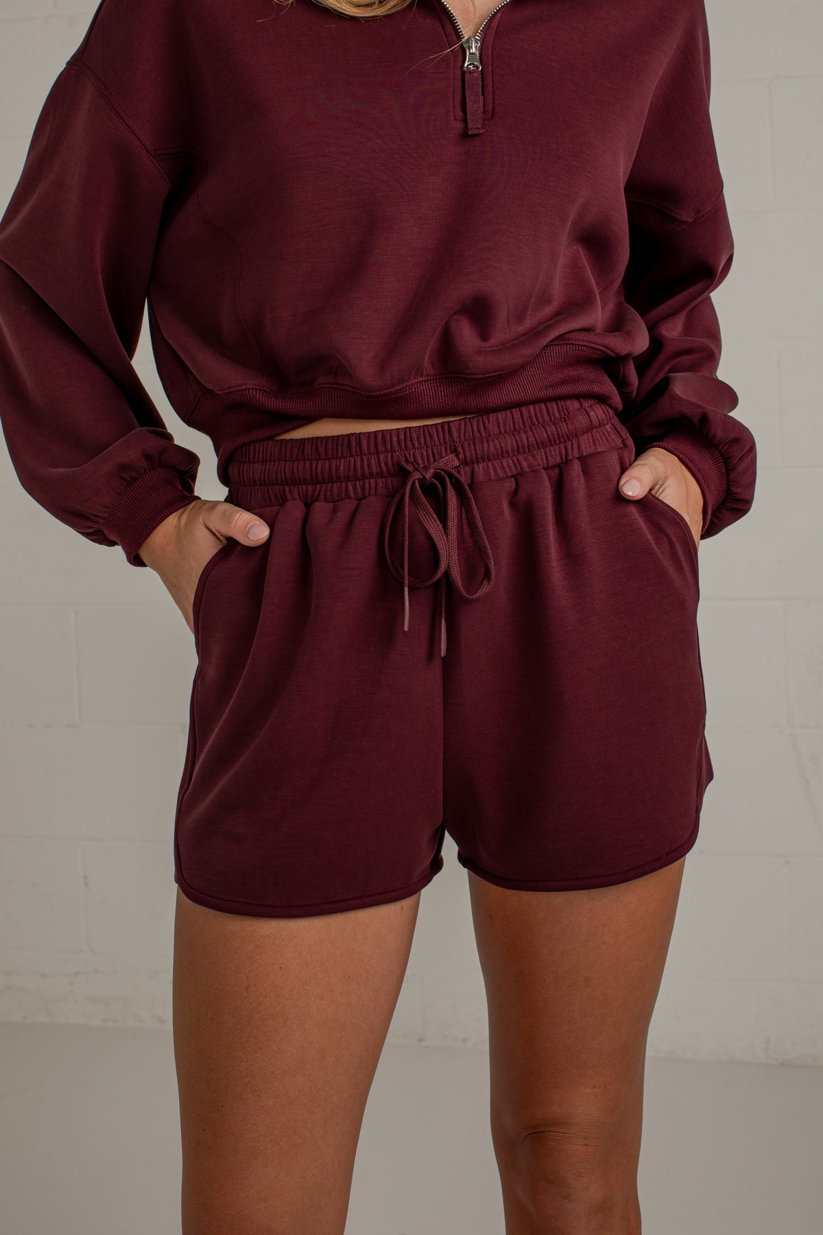 A person wearing a matching burgundy lounge set consisting of a cropped half-zip long-sleeve top and drawstring shorts, standing against a light gray brick wall.