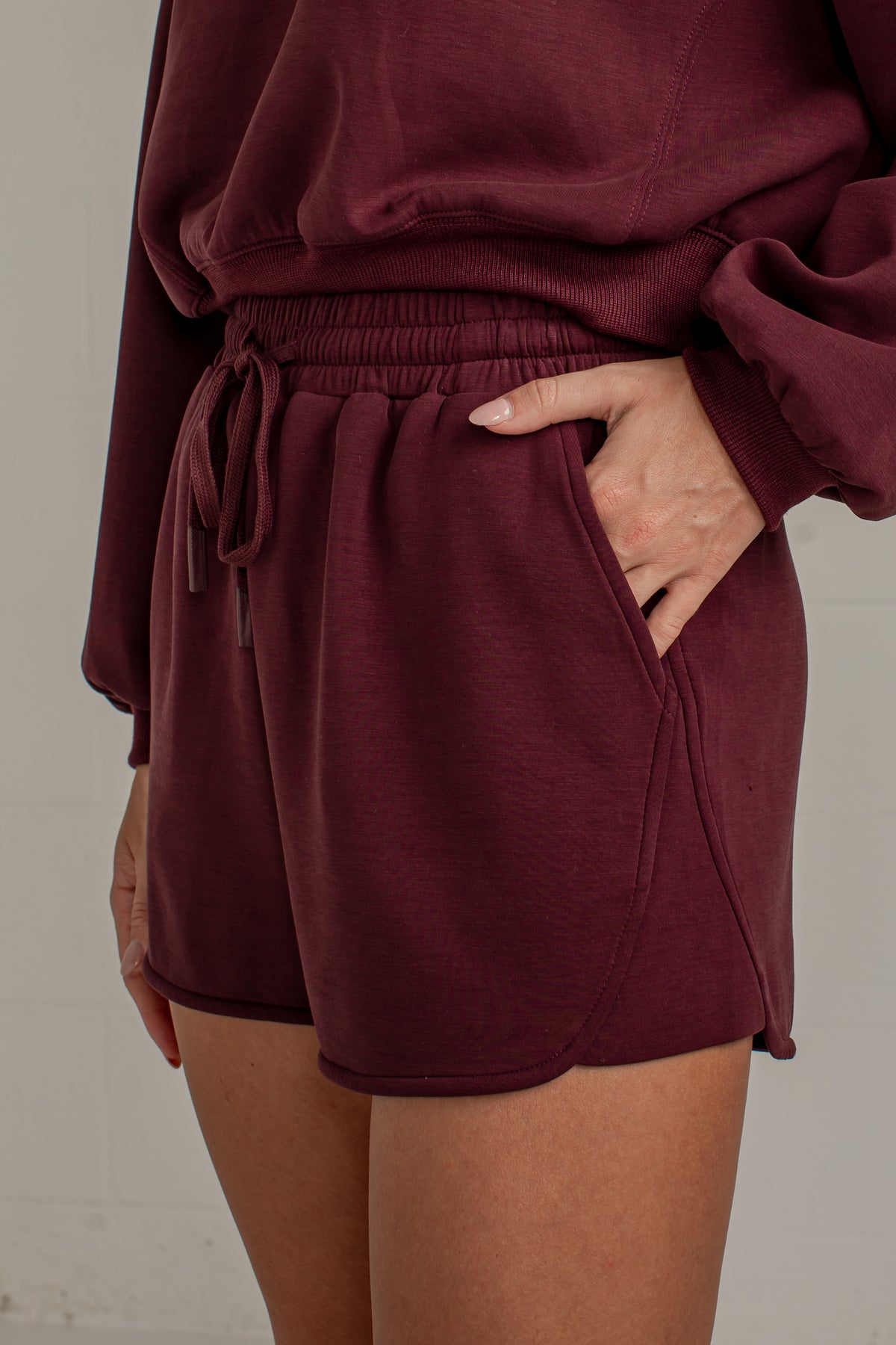 A close-up of a person wearing burgundy drawstring shorts and a matching sweatshirt, with one hand in the pocket of the shorts against a light neutral wall.