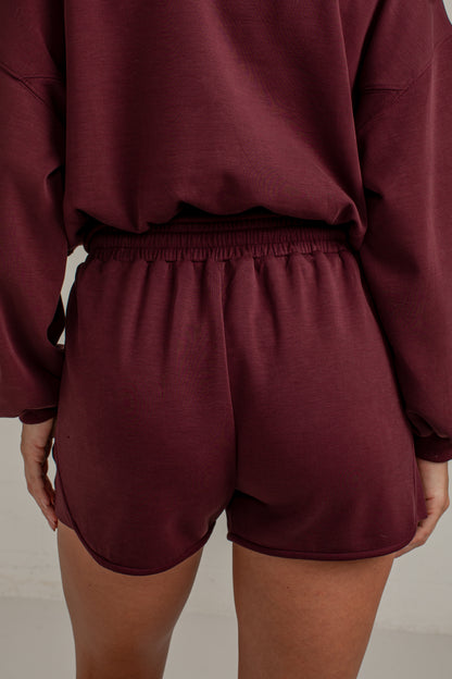 A close-up back view of a person wearing a matching burgundy lounge set, featuring a long-sleeve top and high-waisted shorts with an elastic waistband.
