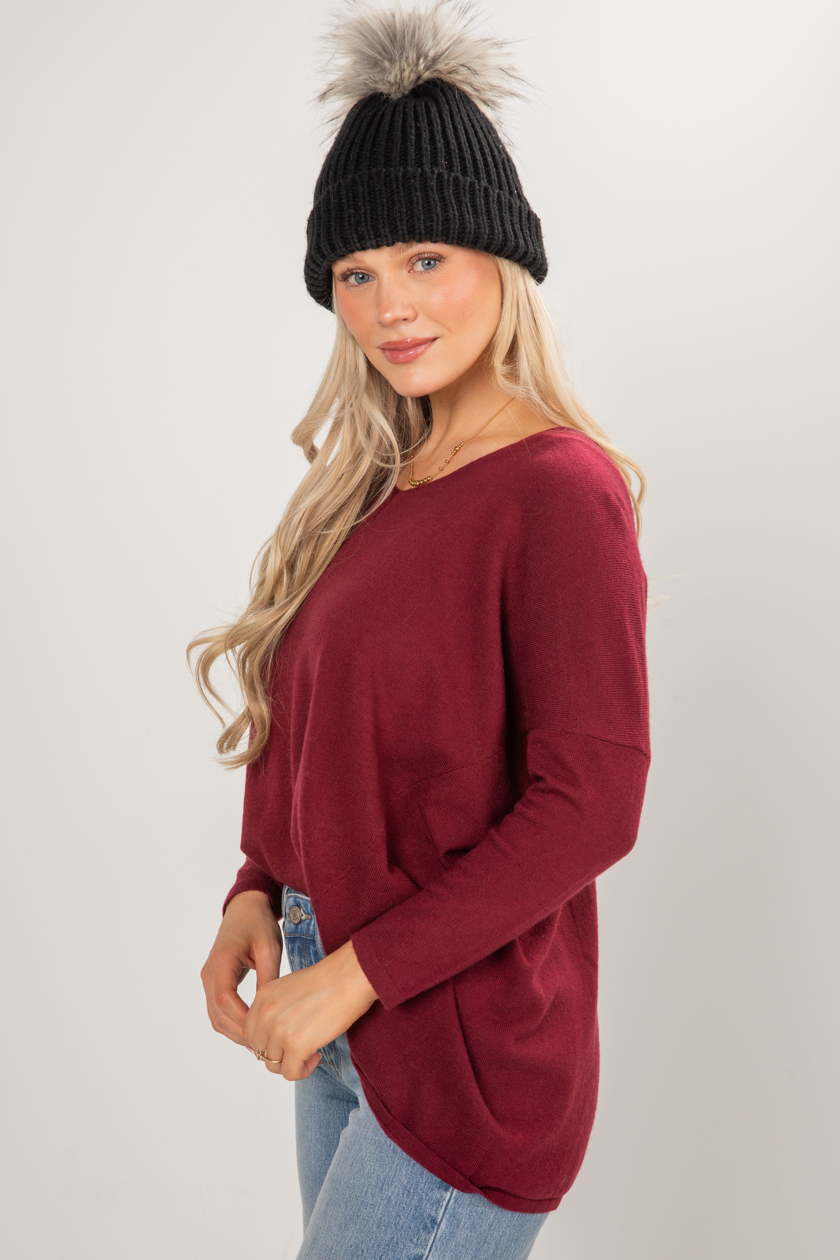 Woman wearing a burgundy sweater and black knit beanie with a fur pom-pom on a white background