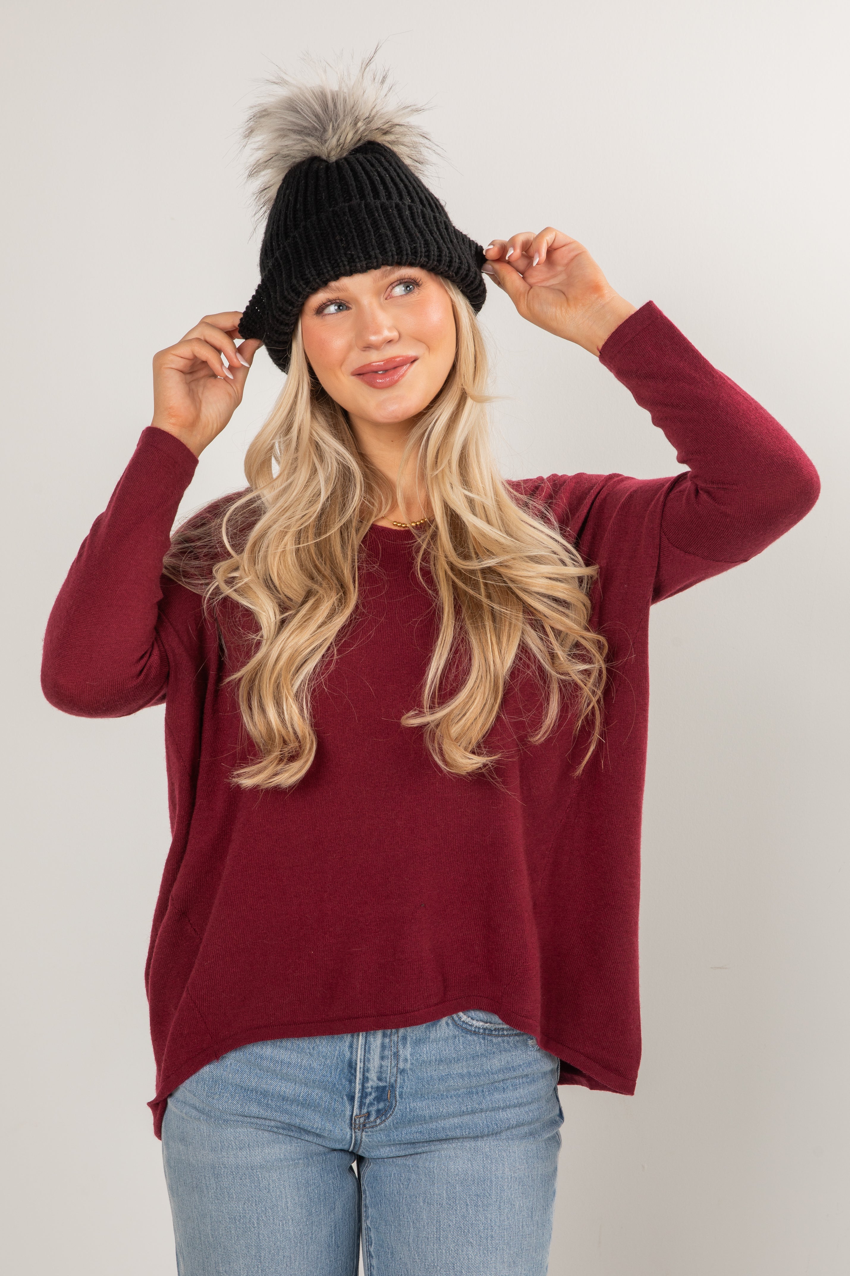 Woman wearing a maroon sweater, black beanie with fur pom-pom, and blue jeans against a white background