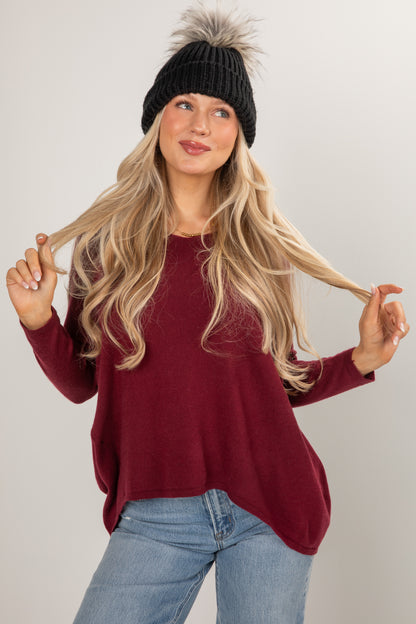 Woman wearing a burgundy sweater and black beanie with fur pom-pom on a plain background
