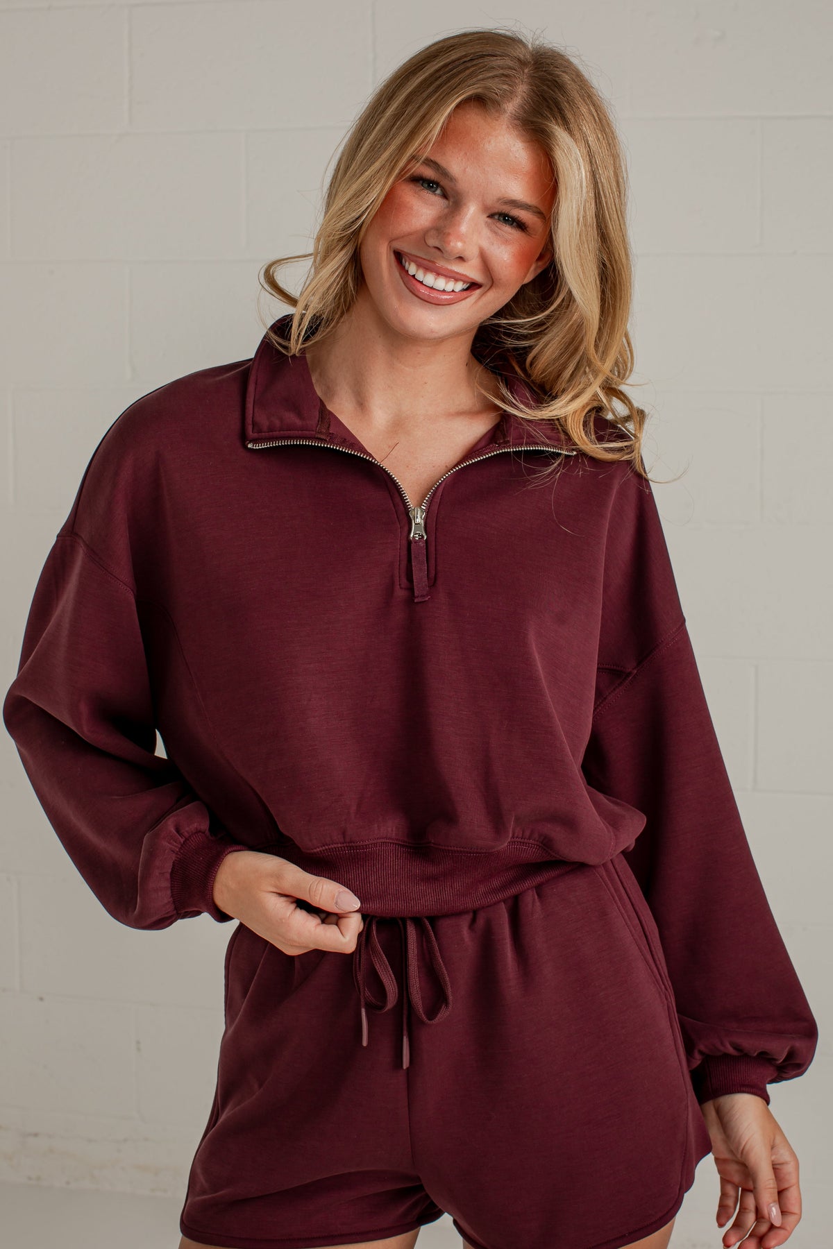 A smiling blonde woman wearing a matching burgundy lounge set consisting of a long-sleeve half-zip sweatshirt and drawstring shorts, standing against a light gray brick wall.