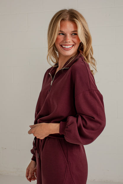 A smiling blonde woman wearing a relaxed-fit maroon zip-up sweatshirt with a matching tie belt, standing against a light gray brick wall.