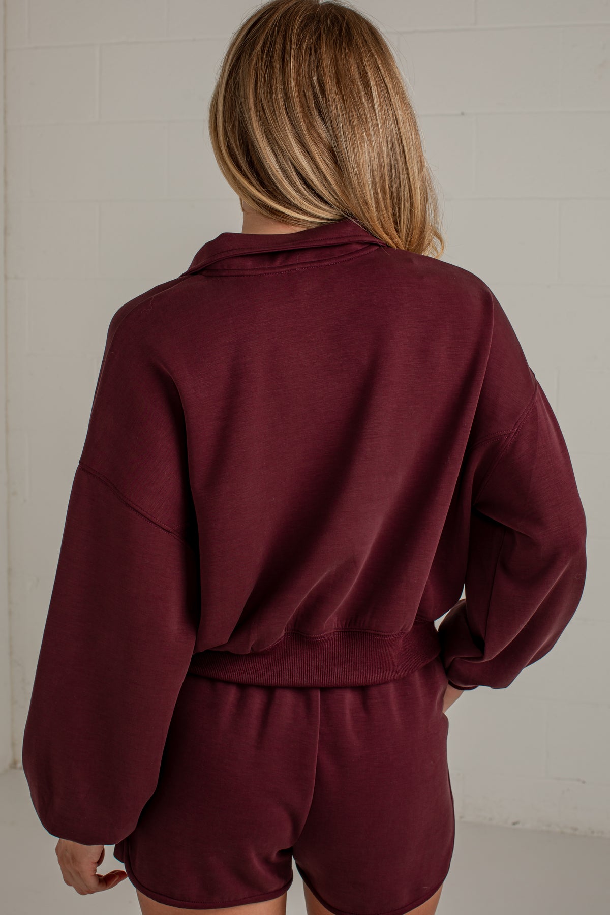 A woman viewed from behind wearing a matching burgundy lounge set consisting of a cropped long-sleeve sweatshirt and shorts, set against a light gray brick wall.