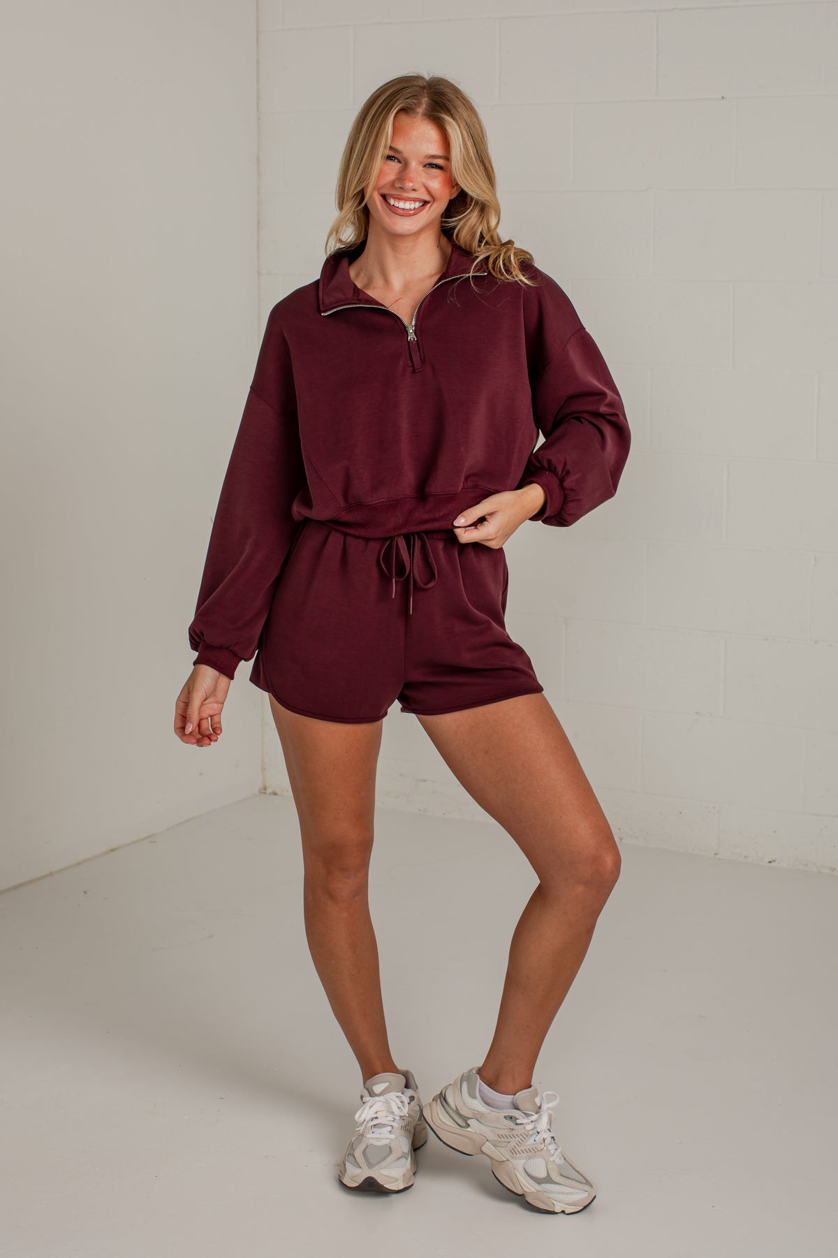 A woman modeling a matching burgundy lounge set consisting of a half-zip long-sleeve top and drawstring shorts, paired with white sneakers, in a studio setting.
