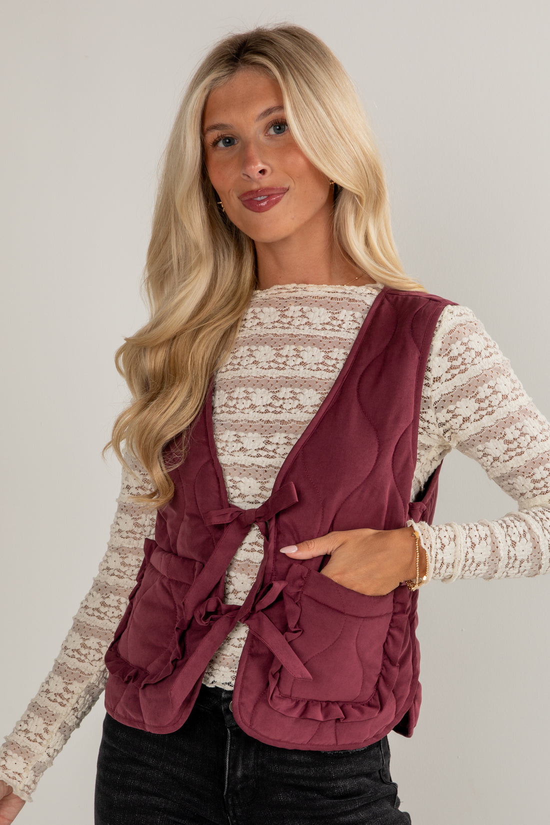 Woman wearing a burgundy vest over a white lace top on a plain background