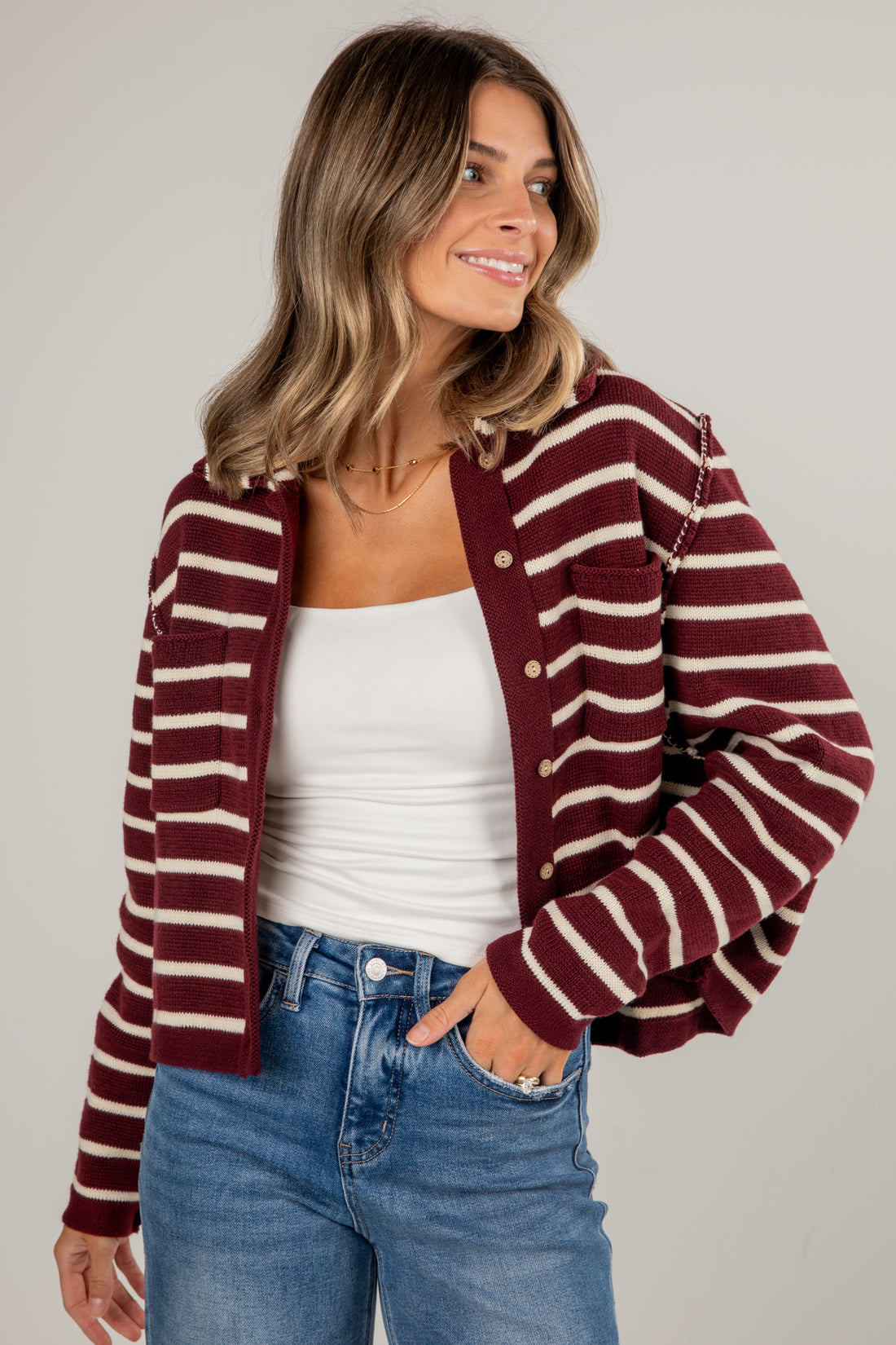 Woman wearing a maroon and beige striped cardigan over a white top and blue jeans on a plain background