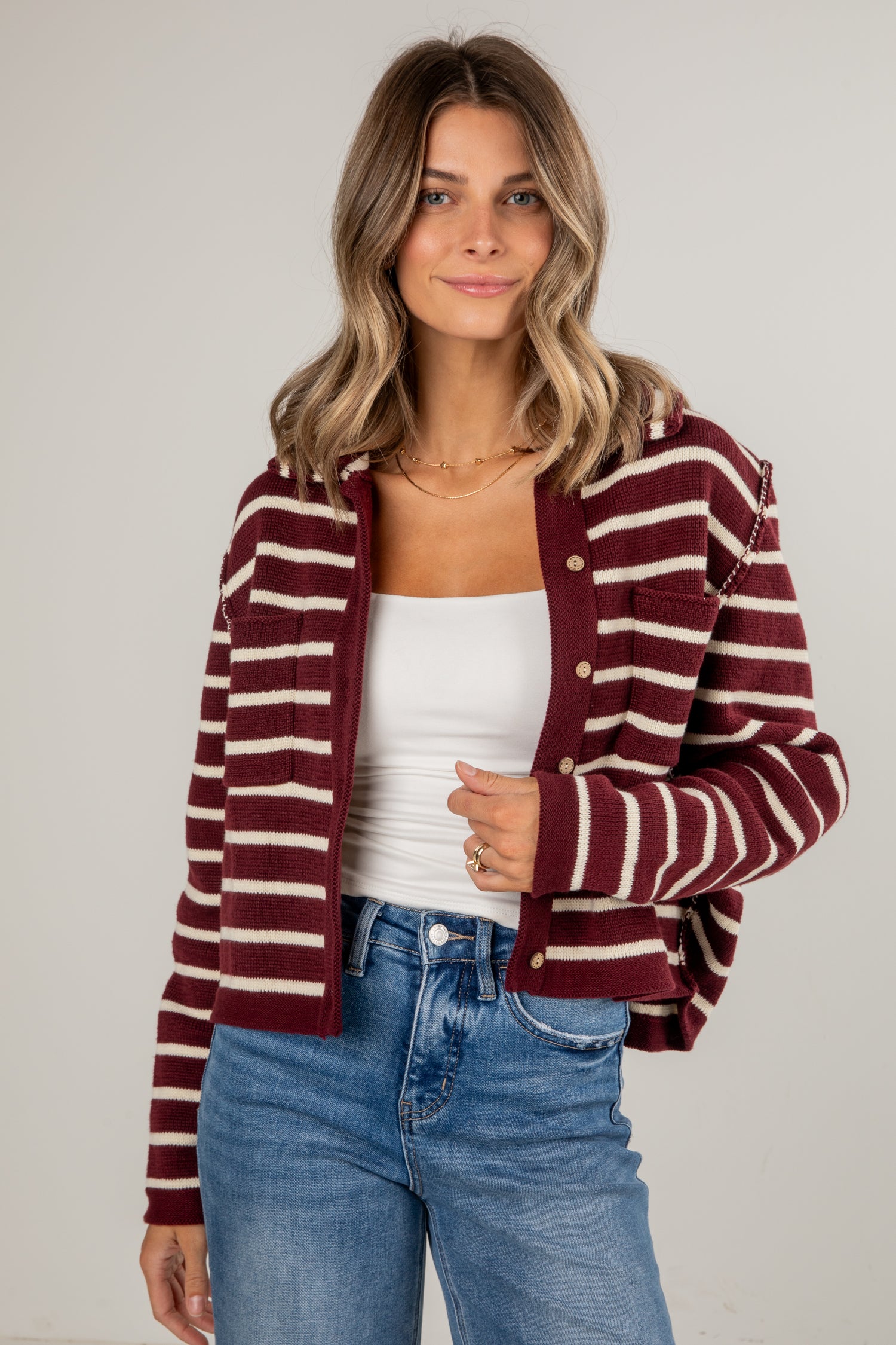 Woman wearing a maroon and white striped cardigan over a white top and blue jeans on a plain background