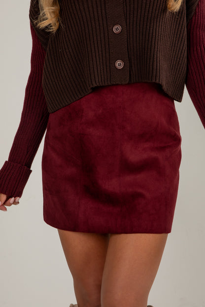 Burgundy suede skirt worn with a brown ribbed sweater on a neutral background